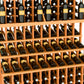 Light wood triple-tier wine bottle display rack with double-deep base, filled with dark/amber bottles